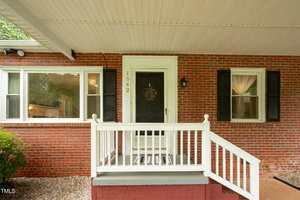 A brick house with two windows, a black front door with a wreath, and the address number 1542. White railing and steps lead up to the small covered porch. Black shutters frame the right window.