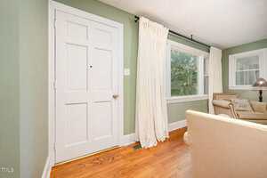 A white door on a light green wall is next to a window with white curtains. Hardwood floors and beige armchairs are visible, along with a side table and lamp by another window in the living room.