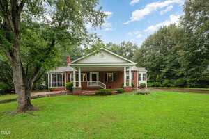 A single-story brick house with a covered front porch, white columns, and large windows sits on a well-maintained lawn, surrounded by tall trees and a curved driveway under a partly cloudy sky.