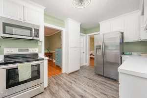 A modern kitchen with white cabinets, stainless steel appliances, a green accent wall, and light gray wood flooring. An oven and microwave are on the left, with a refrigerator on the right and a towel hanging on the oven handle.