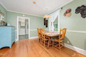 A dining room with light green walls, a wooden table set for four, and matching wooden chairs. A window with white curtains lets in natural light. A blue sideboard and wall art are visible, with hardwood flooring throughout.