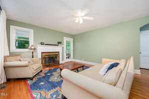 A living room with green walls, hardwood flooring, a fireplace with a wooden mantel, a beige sofa, an armchair, a wooden coffee table, a colorful rug, and a ceiling fan with lights.
