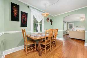 A dining area with a wooden table set for four, wooden chairs, green walls, artwork, and a window with white curtains. An arched doorway leads to a living room with wood floors and a sofa.