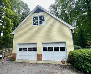 A two-car garage with yellow siding, two white garage doors, and a windowed upstairs area, surrounded by trees and greenery. There are a few flower pots near the right garage door.
