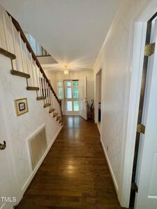 A hallway with hardwood floors, white walls, and a wooden staircase on the left. A light fixture hangs from the ceiling, and double doors at the end open to a bright room. An open door is on the right.