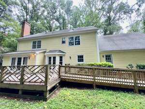 Two-story yellow house with white trim, a shingled roof, brick chimney, and several windows. A wooden deck with a ramp extends from the back, surrounded by grass and trees.