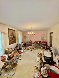 A living room with cream carpeting and pink walls, filled with various household items, boxes, and decorations scattered and stacked throughout the space. A chandelier hangs from the ceiling above.