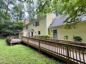 A yellow two-story house with white trim and a sloped roof, featuring a long wooden ramp leading to a deck, surrounded by grass and trees.