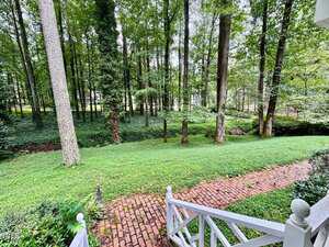 View from a porch showing a brick walkway, grass lawn, and numerous tall trees in a wooded area. A white wooden railing borders the porch, and dense green foliage fills the background.