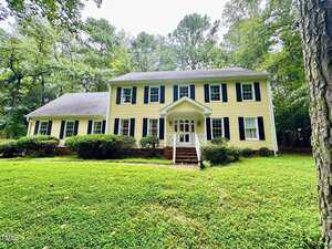 A two-story yellow house with white trim and black shutters at 1744 Parker Lane, Henderson, featuring a central white-columned porch, surrounded by shrubs and grass, with tall trees in the background.