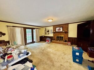 A living room with beige carpet, brick fireplace, wall-mounted shelves, a closed TV cabinet, stacked blue storage bins, a table with assorted items, and French doors with curtains leading outside.