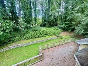 View from an elevated deck overlooking a grassy backyard bordered by a stone retaining wall and dense, leafy trees and shrubs. A wooden railing runs along the edge of the deck.