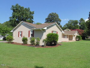 Single-story beige brick house with brown shingles, white trim, and red shutters, surrounded by green grass, shrubs, and trees under a clear blue sky.