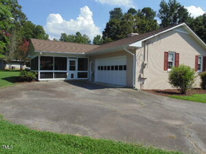 Single-story house with a brick exterior, red shutters, and an attached two-car garage. A screened-in porch is on the left, and the house is surrounded by grass, shrubs, and trees. The driveway is paved.