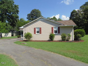 Single-story beige house with a brown shingle roof, red shutters, and white trim. The front yard has green grass, bushes, and a curved driveway. Tall trees are in the background under a blue sky with a few clouds.