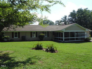 Single-story house with light-colored exterior, dark shutters, and a screened-in porch on the right side, set on a grassy lawn with shrubs and mature trees in the background.