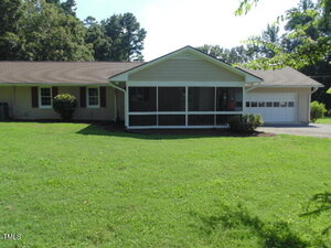 Single-story house with a screened front porch, attached garage, tan exterior, brown roof, and large green lawn. There are bushes near the house and trees in the background.