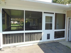 A screened-in porch with a white-framed door and large windows. The porch has dark railing, a ceiling fan inside, and a view of the yard through the screens. The ground outside is concrete.