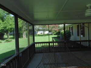 View from a screened-in porch with a ceiling fan and hammock, looking out onto a green backyard with trees, shrubs, and a neighboring house visible in the background.