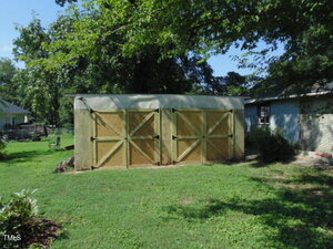 A wooden shed with two large double doors stands in a grassy backyard, shaded by a tree. A blue house is partially visible to the right. The sky is clear and sunny.