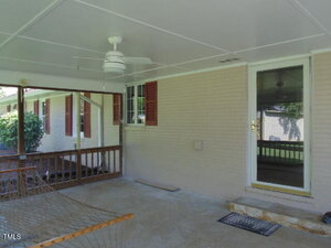 A covered patio with a ceiling fan, wooden railing, and a glass door leading inside. A window with red shutters is on the brick wall, and a hammock is partially visible in the foreground.