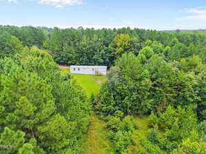 A white mobile home sits in a clearing surrounded by dense green trees and grassy areas under a blue sky with scattered clouds.