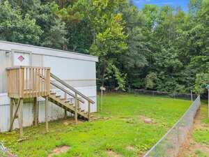 A white mobile home with a small wooden staircase and porch sits beside a fenced grassy yard, surrounded by dense, green trees under a blue sky.