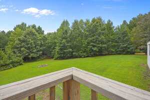 View from a wooden deck railing overlooking a large green lawn bordered by dense trees, with a fire pit area visible in the grass and a small portion of a building on the right edge. Blue sky with scattered clouds above.