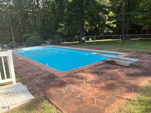 Rectangular outdoor swimming pool with clear blue water at 80 White Deer Lane, Henderson, surrounded by a reddish stone-patterned deck. Features a diving board on one end, a white fence, and trees and grass in the background.