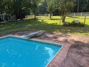 A backyard with a rectangular swimming pool, diving board, and stone-patterned pool deck. Beyond the chain-link fence is a grassy yard with trees and a white wooden fence.