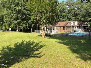 A brick house with dormer windows is seen in the background, with a fenced-in pool and a grassy yard in the foreground. Trees surround the property, and a basketball hoop stands at the edge of the lawn.