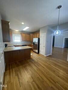 A kitchen with wooden cabinets, stainless steel refrigerator, beige countertops, hardwood floors, and a window above the sink. An adjacent room with hardwood flooring is visible through an open doorway.