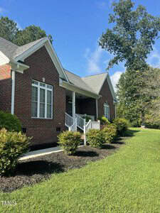A red brick house with white trim and a gray roof, featuring a small front porch with white railings, surrounded by neatly trimmed bushes and a green lawn under a blue sky.