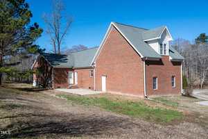 A red brick house with a gray shingle roof is shown on a clear day. The house has several windows, a white door, and a small concrete patio. The yard is sloped with sparse grass and some trees in the background.
