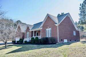 This charming single-story brick house at 32 Valley Way, Henderson, features white trim, a covered front porch, multiple gables, and foundation shrubs. The yard offers grass, a small tree, and is bordered by peaceful woods in the background.