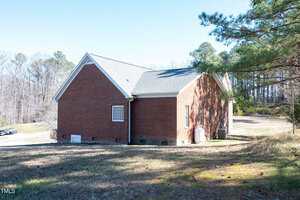 A red brick house with a gray shingled roof sits on a grassy lot, bordered by trees. The house has multiple windows and a propane tank near the side. The sky is clear and sunny.