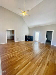 A spacious room with light wood flooring, vaulted ceiling, ceiling fan, a fireplace in the corner, and doorways leading to adjacent rooms; a windowed door is on one wall.