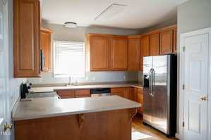 A kitchen with light brown wooden cabinets, beige countertops, a stainless steel refrigerator, built-in microwave, dishwasher, electric stove, and a window above the sink allowing natural light in.