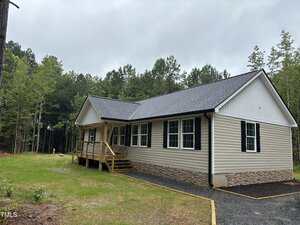 A single-story beige house with black shutters and a dark gray roof sits on a grassy lot bordered by trees. A small covered porch with wooden steps leads to the front door. The sky is overcast.