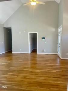 A spacious, empty room with light hardwood floors, gray walls, a ceiling fan with light, vaulted ceilings, and a door leading to another room. A thermostat and vent are visible on one wall.