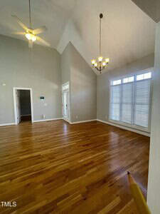 A spacious empty room with light gray walls, hardwood floors, a ceiling fan, chandelier, high vaulted ceiling, large window with blinds, and a small doorway visible in the background.