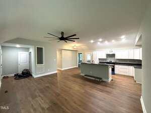 Open-concept living area with wood flooring, ceiling fan, and recessed lighting. The modern kitchen features white cabinets, stainless steel appliances, and a large island. Some tools and equipment are on the floor near the entryway.
