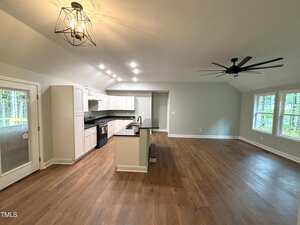 Open-concept kitchen and living area with hardwood floors, white cabinets, black countertops, recessed and pendant lighting, island with sink, large windows, and ceiling fans. Glass door leads outside. Room is unfurnished.