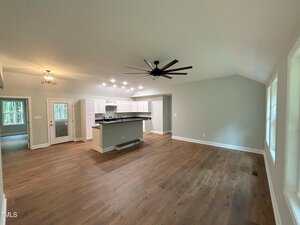 A spacious open-concept room with wood flooring, white kitchen cabinets, a large black ceiling fan, multiple ceiling lights, and several windows letting in natural light. A kitchen island separates the kitchen from the living area.