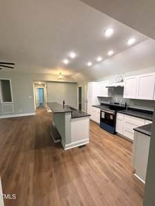 A modern kitchen with white cabinets, black countertops, an island in the center, and wood-style flooring. Recessed and pendant lighting brighten the space. A hallway and additional rooms are visible in the background.