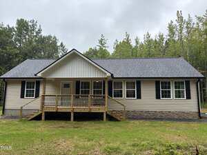 A single-story beige house with black shutters, a front porch with wooden railings, and a dark shingled roof sits on 2665 Bodie Currin Road in Oxford, surrounded by grass and trees on a cloudy day.