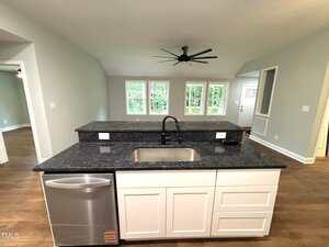 A modern kitchen island with a dark countertop, built-in sink, dishwasher, and white cabinets faces a living area with large windows, light walls, wood flooring, ceiling fan, and a glimpse of the front door.