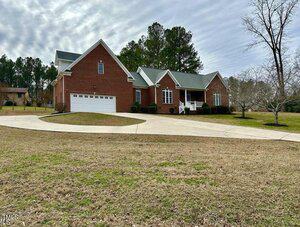 A large brick house with a green roof, white trim, and an attached three-car garage. The house has a circular driveway and sits on a spacious lawn with a few trees and shrubs.