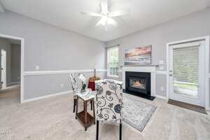 A small dining area with two patterned chairs and a table on beige carpet is set near a gas fireplace. A window and glass door let in natural light. The walls are light gray with white trim and a ceiling fan above.