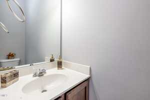 A bathroom with a white sink, chrome faucet, soap dispenser, and toothbrush holder on a beige countertop. A large mirror reflects part of the sink and a toilet with a small plant decoration.